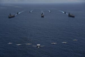 Aircraft from Carrier Air Wing (CVW) 9 fly over the Nimitz-class aircraft carrier Abraham Lincoln (CVN-72), front left, America-class amphibious assault ship USS Tripoli (LHA-7), front center, Nimitz-class aircraft carrier USS Ronald Reagan (CVN-76), front right, Ticonderoga-class guided-missile cruiser USS Mobile Bay (CG -3), middle left, Arleigh Burke-class guided-missile destroyer USS Benfold (DDG-65), middle center, Ticonderoga-class guided-missile cruiser USS Antietam (CG-54), middle right, Arleigh Burke-class guided-missile destroyer USS Spruance (DDG-111), back left, and Arleigh Burke-class guided-missile destroyer USS Fitzgerald (DDG-62), back right, as they sail in formation during the biennial field training exercise Valiant Shield 2022 in the Philippine Sea. (Photo: U.S. Navy by Mass Communication Specialist 3rd Class Thaddeus Berry)