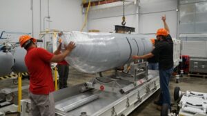 From left: Kennie Martinez and Marc Dannemiller, Raytheon Intelligence & Space employees, unbox the first of two Next Generation Jammer Mid-Band fleet representative pods that were delivered to the Airborne Electronic Attack Systems (PMA-234) pod shop at Naval Air Warfare Center Aircraft Division, Patuxent River, Md. on July 7, 2022. (Photo: U.S. Navy)